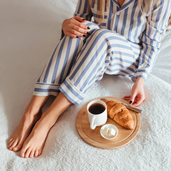 Γυναίκες όμορφα πόδια σε πιτζάμες και δίπλα σε έναν καφέ με ένα κρουασάντ.την έννοια του καφέ στο κρεβάτι — Φωτογραφία Αρχείου