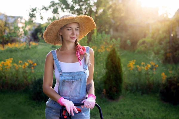 Tukang kebun wanita. Seorang gadis seksi dengan topi memotong rumput dekat rumah dengan mesin pemotong rumput. Desain lansekap dan perawatan halaman — Stok Foto
