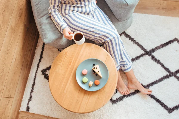 Una mujer en pijama por la mañana en una mesa bebe café y come dulces tarta de queso macarrones dulces. el concepto de desayuno con dulces —  Fotos de Stock