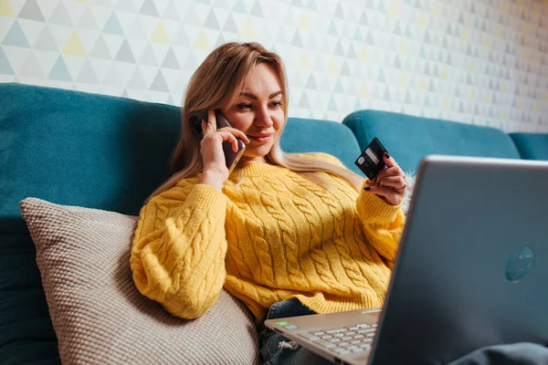 Een vrouw met een laptop in een gele trui zit op de bank en rijdt met een bepaalde creditcard. het concept van kopen terwijl je thuis zit — Stockfoto