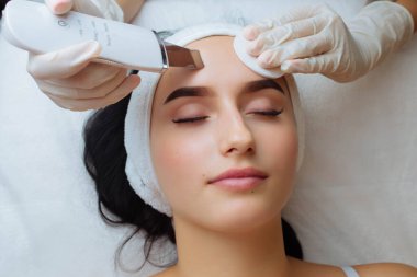 Cildi ultrason fırçasıyla temizleme işlemi sırasında güzel beyaz bir bayanın üst görüntüsünü kapatın. Profesyonel kadın afro güzellik uzmanının elinde ultrasonik cihaz var.