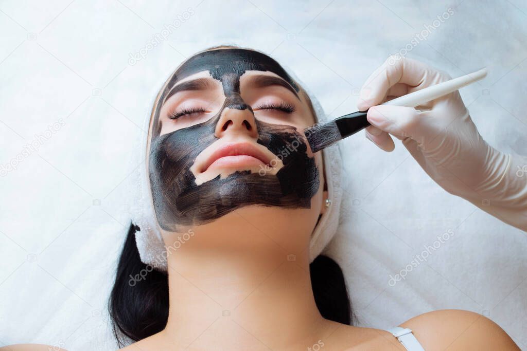cosmetologist applying black mask on pretty woman face wearing black gloves, gorgeous woman in spa having facial procedures.