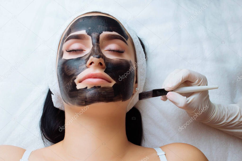 cosmetologist applying black mask on pretty woman face wearing black gloves, gorgeous woman in spa having facial procedures.