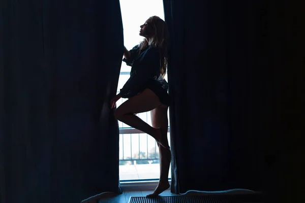 自宅でストリップ。窓の前でセクシーなブロンドがドレスアップ。魅力的な女の子のシルエット — ストック写真