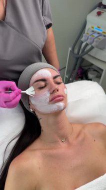 Estetikçi cilt bakımı sırasında fırça kullanan bir kadının yüzüne beyaz yüz maskesi takar.