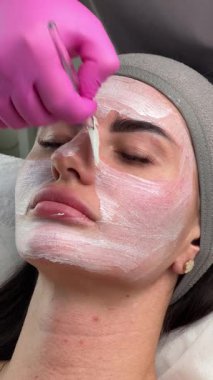 Estetikçi cilt bakımı sırasında fırça kullanan bir kadının yüzüne beyaz yüz maskesi takar.