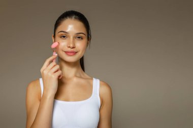Yanağında pembe bir yüz silindiri kullanan genç bir kadın yumuşak bir stüdyo arka planında rahatlatıcı bir cilt masajı sergiliyor.