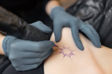 Dövmeci bir müşterinin cildine güneş tasarımı çizerken, dövme sürecinin hassasiyetini, steril eldivenleri ve profesyonel ekipmanları gösterir.