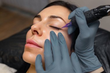 Bir kozmetik uzmanının göz kapağı makyajı, göz kalemi dövme prosedürünü steril aletlerle gerçekleştirmesi profesyonel bir stüdyoda kırbaç çizgisini ve göz güzelliğini artırması.