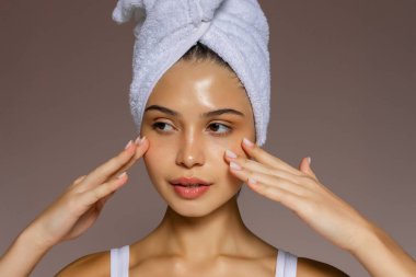 Duştan sonra iki eliyle yüzüne masaj yapan, kafasına havlu takan ve temiz bir cilt göstergesi olan genç bir kadın.