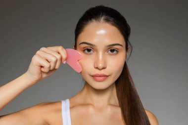 Genç bir kadın yüzüne pembe bir gua sha taşıyla nazikçe masaj yapıyor. Tarafsız bir arkaplana karşı cilt bakımını gösteriyor.