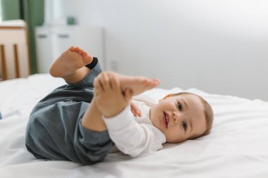 Yumuşak beyaz bir yatakta dinlenen neşeli bir bebek, sıcak ve rahat bir ev anının tadını çıkarıyor. Hafif ışık, rahatlık, masumiyet ve doğal yaşam tarzı atmosferiyle.