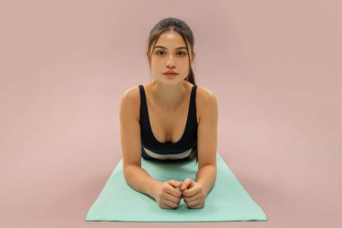 Egzersiz sırasında yoga minderinde uzanan genç bir kadın pastel arka planda fitness tahtası varyasyonu üzerinde çalışırken ileriye bakıyor.