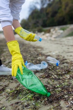 Kirli bir plajda plastik şişe toplamaya gönüllü olan gönüllüler, okyanus kirliliği, atık sorunu, çevre koruma ve kıyı temizliği eylemlerini vurguluyor