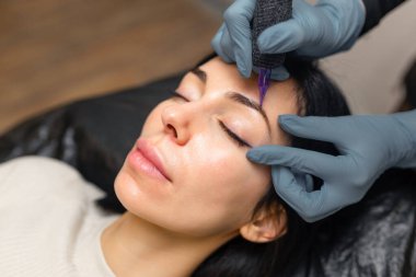 Kadın bir müşteriye kaş mikroplu pateni yapan bir kozmetikçiye yakın çekim. Profesyonel güzellik prosedürü, steril aletler, hassas işler ve modern bir güzellik stüdyosunda kaş şekillendirme.