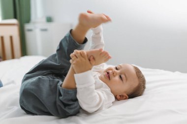 Yumuşak beyaz bir yatakta dinlenen neşeli bir bebek, sıcak ve rahat bir ev anının tadını çıkarıyor. Hafif ışık, rahatlık, masumiyet ve doğal yaşam tarzı atmosferiyle.