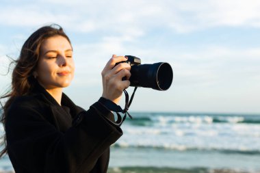Güneşli bir plajda elinde kamerayla gülümseyen kadın fotoğrafçı, altın saatin tadını çıkarıyor ve sahildeki açık hava anlarını yakalıyor.