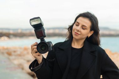 Genç bayan fotoğrafçı deniz kenarında fotoğraf çekiyor, elinde flaşla profesyonel bir kamera tutarken gülümsüyor, gün batımında kayalık bir sahil şeridinde anları yakalıyor.
