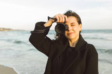 Güneşli bir plajda elinde kamerayla gülümseyen kadın fotoğrafçı, altın saatin tadını çıkarıyor ve sahildeki açık hava anlarını yakalıyor.