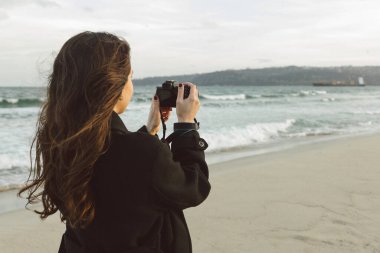 Güneşli bir plajda elinde kamerayla gülümseyen kadın fotoğrafçı, altın saatin tadını çıkarıyor ve sahildeki açık hava anlarını yakalıyor.