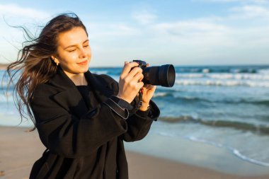 Güneşli bir plajda elinde kamerayla gülümseyen kadın fotoğrafçı, altın saatin tadını çıkarıyor ve sahildeki açık hava anlarını yakalıyor.