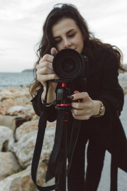 Kadın fotoğrafçı, deniz kenarındaki tripoda profesyonel kamerayı ayarlıyor. Kayalık bir sahil şeridinde yaratıcı bir fotoğraf çekimine hazırlanırken objektife odaklanıyor.