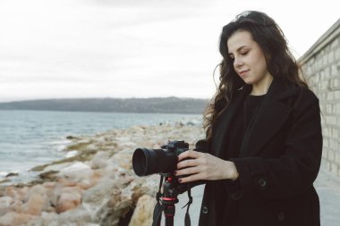Kadın fotoğrafçı, deniz kenarındaki tripoda profesyonel kamerayı ayarlıyor. Kayalık bir sahil şeridinde yaratıcı bir fotoğraf çekimine hazırlanırken objektife odaklanıyor.
