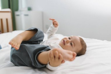 Yumuşak beyaz bir yatakta dinlenen neşeli bir bebek, sıcak ve rahat bir ev anının tadını çıkarıyor. Hafif ışık, rahatlık, masumiyet ve doğal yaşam tarzı atmosferiyle.