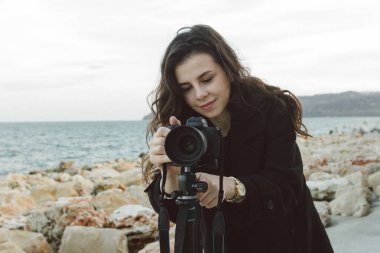 Kadın fotoğrafçı, deniz kenarındaki tripoda profesyonel kamerayı ayarlıyor. Kayalık bir sahil şeridinde yaratıcı bir fotoğraf çekimine hazırlanırken objektife odaklanıyor.