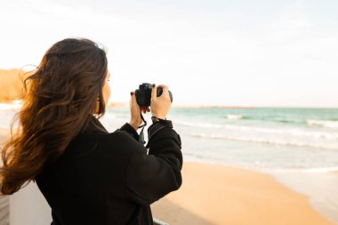 Güneşli bir plajda elinde kamerayla gülümseyen kadın fotoğrafçı, altın saatin tadını çıkarıyor ve sahildeki açık hava anlarını yakalıyor.
