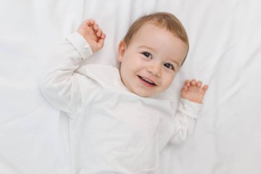 Evdeki yumuşak beyaz bir yatakta uzanmış, doğal ışıkta gülümseyen mutlu bir bebeğin samimi portresi. Sıcak ve rahat bir çocukluk anı neşe, masumiyet, rahatlık ve nazik aile duygularıyla dolu.