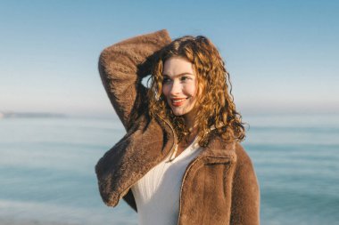 Gün doğumunda gölün kenarında huzurlu bir anın tadını çıkaran genç bir kadın. Kıvırcık saçlar, sıcak güneş ışığı, doğal güzellik ve sakin yaşam tarzı, yumuşak ışık, özgürlük ve rahatlama kavramı.