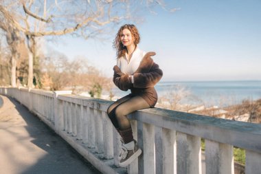 Deniz kenarındaki korkuluklarda oturan, güneşli deniz gününün tadını çıkaran genç bir kadın. Günlük kıyafet, rahat poz, sahil şehri yaşam tarzı, özgürlük ve sakin açık hava atmosferi.