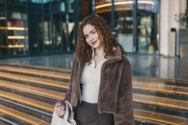 Şehirde modern cam binanın yakınında poz veren genç bir kadın. Şık günlük kıyafet, kendine güvenen görünüş, çağdaş şehir yaşam tarzı, akşam ışığı ve şık atmosfer.