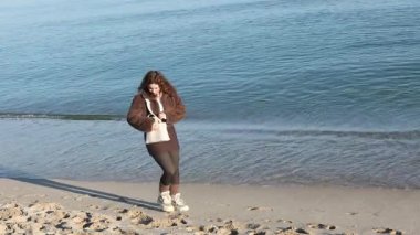 Genç bir kadın deniz kenarındaki kumsalda yürüyor, çantasından akıllı telefonu alıyor. Açık hava yaşam sahnesi, deniz kenarında sakin bir ortam, doğal ışık, kıyı şeridine yakın her gün. Yüksek kalite 4k