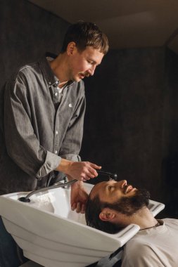 Berber, kuaför lavabosunda şampuanla müşteri saçlarını yıkıyor. Profesyonel erkek bakımı, kafa yıkama, modern berber dükkanında kuaförlük hizmetleri.