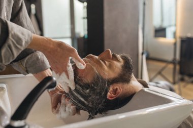 Berber, kuaför lavabosunda şampuanla müşteri saçlarını yıkıyor. Profesyonel erkek bakımı, kafa yıkama, modern berber dükkanında kuaförlük hizmetleri.