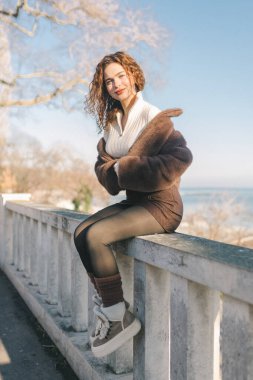 Deniz kenarındaki korkuluklarda oturan, güneşli deniz gününün tadını çıkaran genç bir kadın. Günlük kıyafet, rahat poz, sahil şehri yaşam tarzı, özgürlük ve sakin açık hava atmosferi.