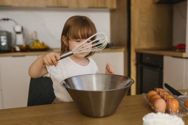 Mutfakta metal kasenin üzerinde çırpı tutan küçük bir kız, evde pişirmeye yardım eden bir çocuk, aile tarzı yemek pişirme sahnesi.