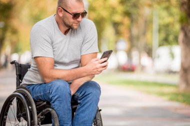 Park sokağındaki tekerlekli sandalyedeki engelli adam akıllı telefon kullanıyor.