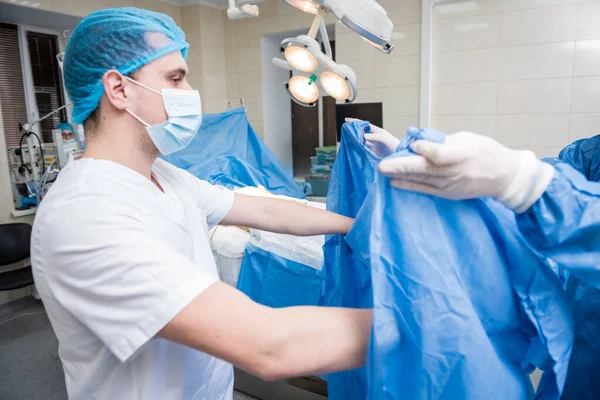 Assistente ajuda o cirurgião a colocar luvas de látex e vestido cirúrgico antes da operação. — Fotografia de Stock