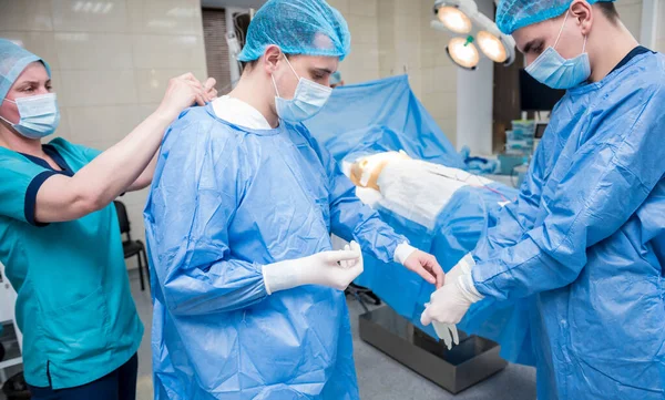 Помощник помогает хирургу надеть латексные перчатки и хирургическое платье перед операцией. — стоковое фото