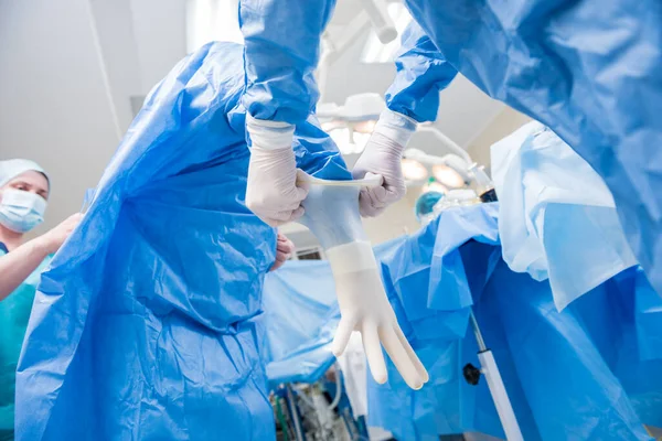 Assistente ajuda o cirurgião a colocar luvas de látex e vestido cirúrgico antes da operação. — Fotografia de Stock