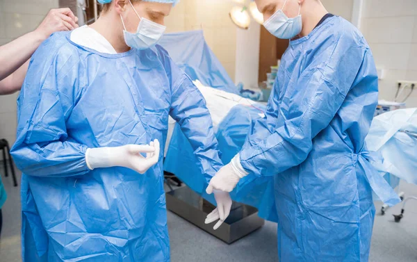 Assistente ajuda o cirurgião a colocar luvas de látex e vestido cirúrgico antes da operação. — Fotografia de Stock