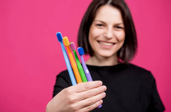 Schöne glückliche junge Frau mit verschiedenen farbigen Zahnbürsten auf leerem rosa Hintergrund — Stockfoto