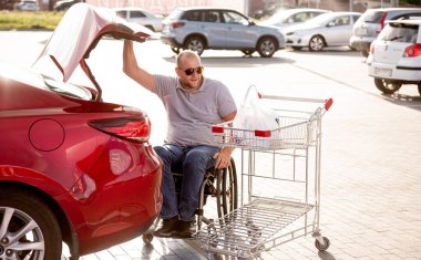 Tekerlekli sandalyedeki yetişkin bir adam süpermarketin otoparkındaki bir arabanın bagajına alışveriş yapıyor.