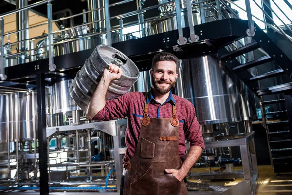 Jonge mannelijke brouwer in lederen schort houdt vat met ambachtelijk bier in moderne brouwerij fabriek — Stockfoto