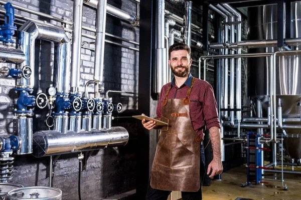 Jovem cervejeiro macho em avental de couro supervisionando o processo de fermentação da cerveja na fábrica moderna de cervejaria — Fotografia de Stock