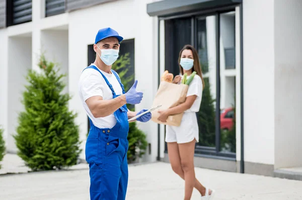 Un joven mensajero con una máscara protectora y guantes entrega mercancías a una joven durante la cuarentena. — Foto de Stock