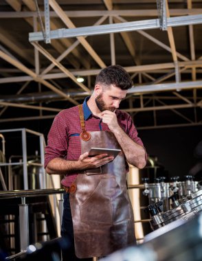 Modern bira fabrikasında bira fermantasyonunu denetleyen deri önlüklü genç bir erkek bira üreticisi.